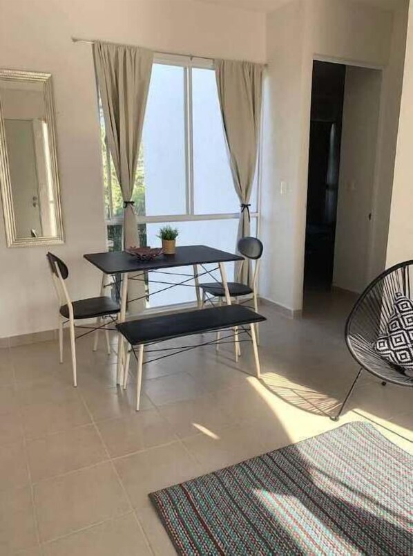A room with a dining table, chairs, a bench, a chair with a patterned cushion, a mirror, and a window with curtains.