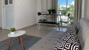 Een moderne woonkamer met een grijze bank, een klein rond tafeltje met een plant en een eethoek met een tafel en stoelen.