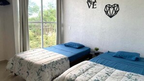 Een kamer met twee bedden, waarvan er een een bloemenprint deken heeft en de ander een effen kleur. Er is een raam met gordijnen, een wand met de tekst 'LOVE' en een geometrisch ontwerp, en een kleine plant op een nachtkastje.