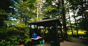 A forested area with a covered outdoor seating area, a table with containers, and a chair. - White Pine Glamping Site (Merville)