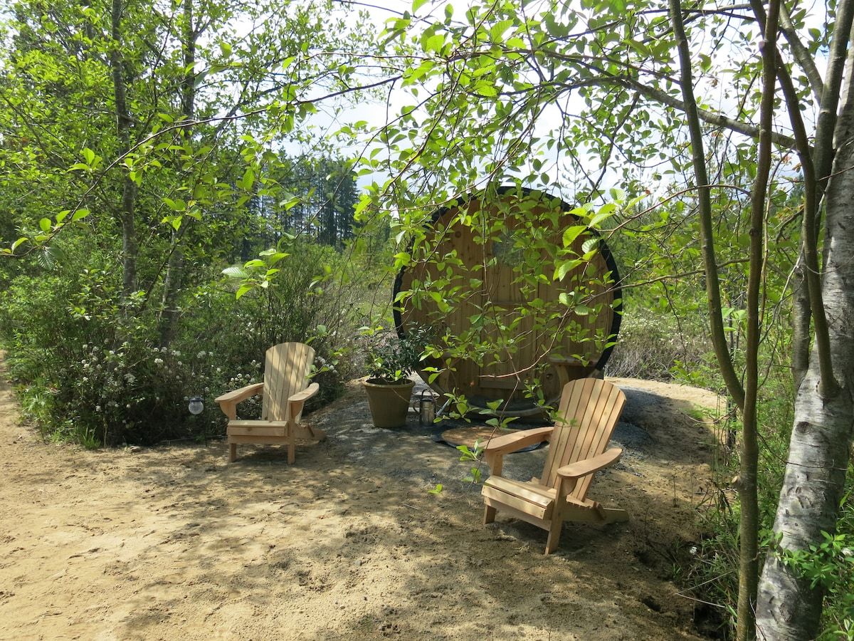 Une terrasse en bois avec deux fauteuils Adirondack, entourée d’une végétation luxuriante et d’un grand tronc d’arbre.