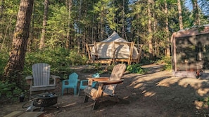A forest campsite with a tent, Adirondack chairs, a portable stove, and a covered structure.