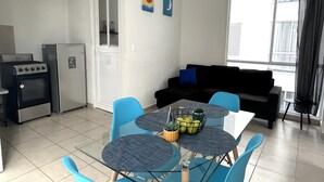 Un espacio habitable compacto con kitchenette, una zona de comedor con sillas azules y una mesa de vidrio, un sofá negro y una ventana con cortinas.