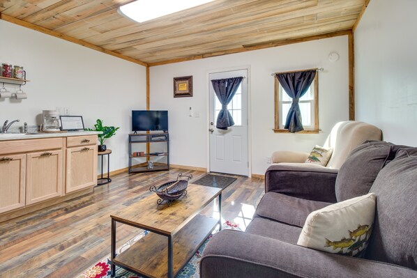 A living room with a wooden ceiling, a sofa, a coffee table, a TV, and a kitchen area with cabinets and a coffee machine.