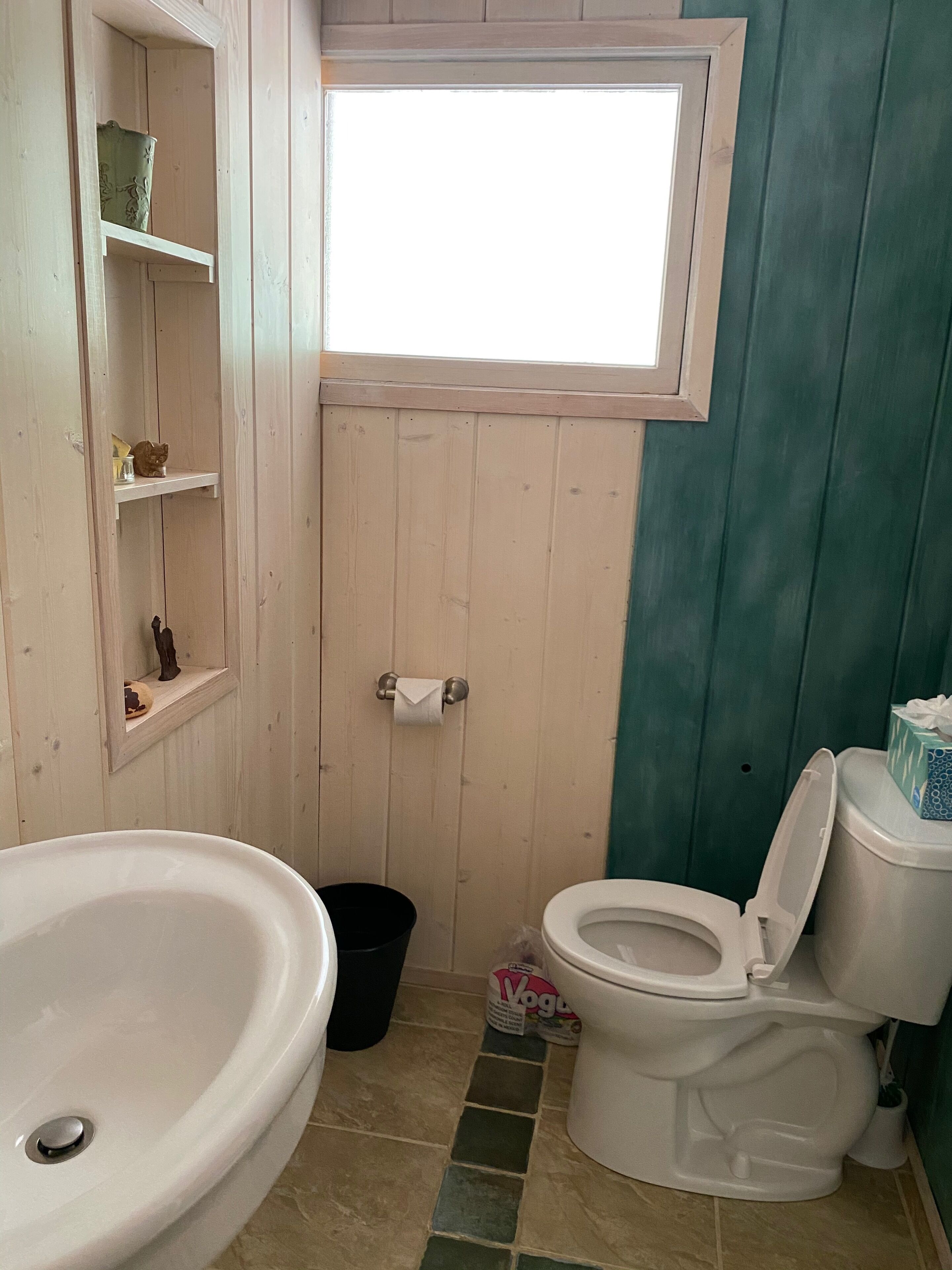 A small bathroom with a white sink, toilet, and a window above the toilet. There are shelves with decorative items and a black trash bin.