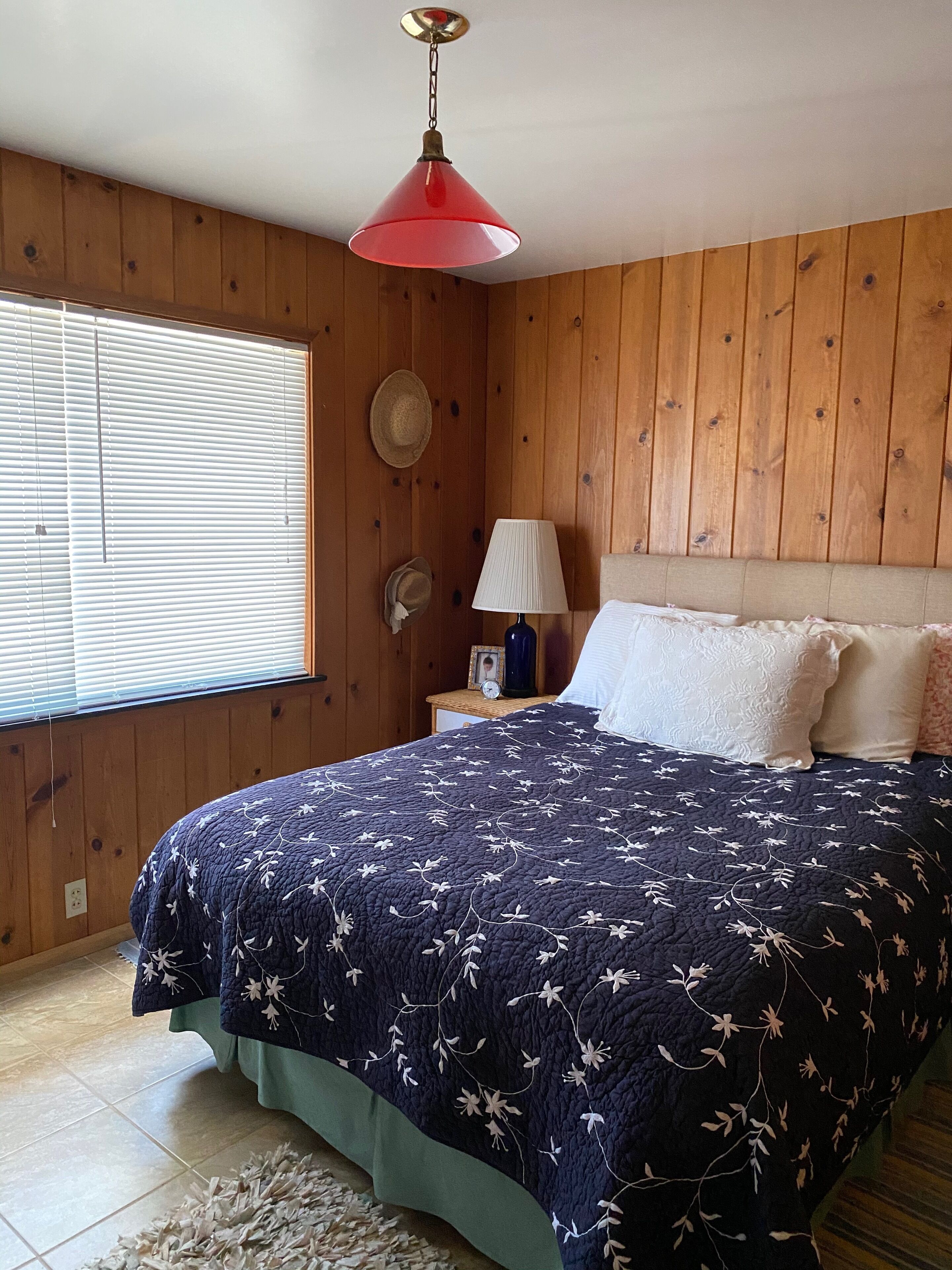 A bedroom with a wooden paneled wall, a bed with a floral quilt, a red hanging lamp, a bedside table with a lamp, and a window with blinds.