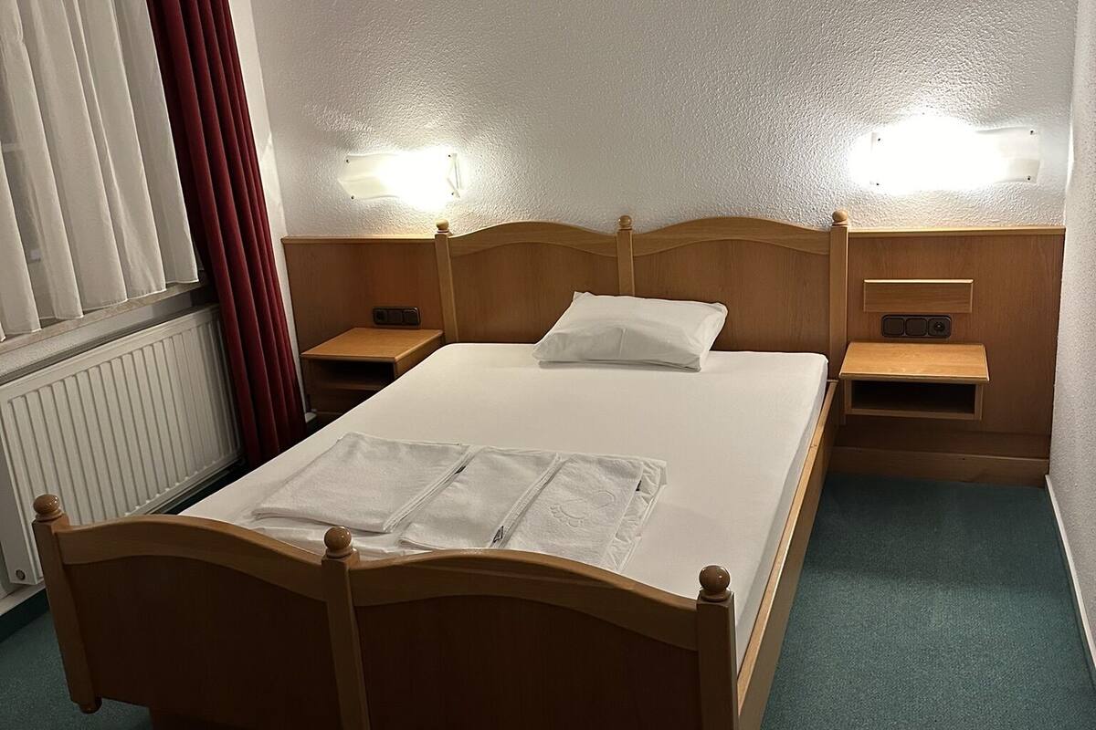 A standard hotel room with a wooden bed, two bedside tables, a nightstand, a radiator, and a window with curtains.