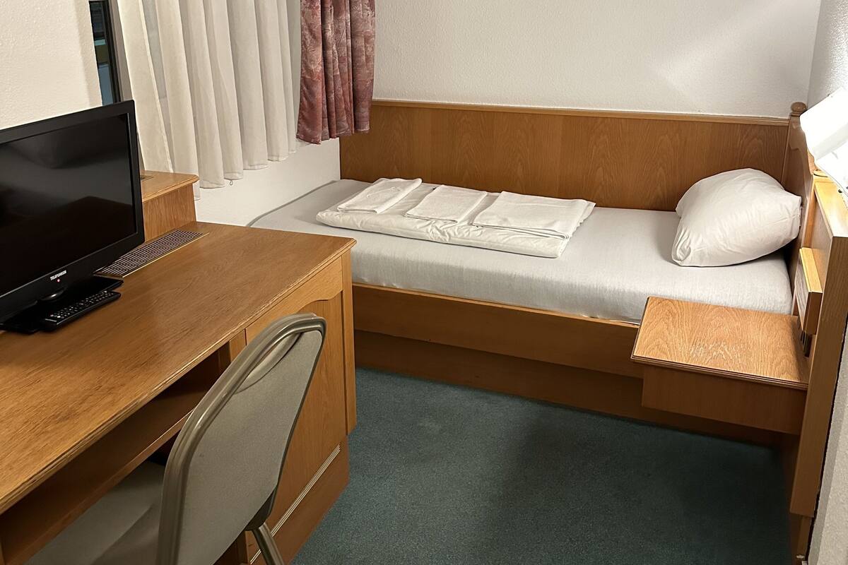 A compact hotel room with a wooden desk, a single bed, and a small chair.