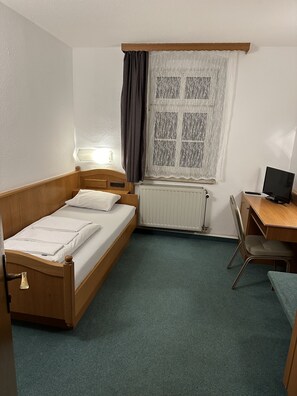 Una habitación pequeña y ordenada con una cama individual, un escritorio, una silla y una ventana con cortinas.