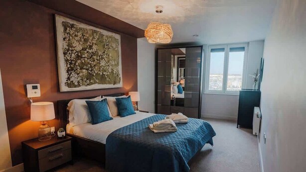 A modern bedroom with a large bed, a nightstand, a wall-mounted light, and a view through an open door. - Marina with a view / 2 bd flat (Brighton and Hove)