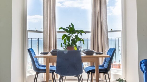 A dining area with a table set for two, blue chairs, a potted plant, and a view of the sea through large windows with curtains. - Sea Breeze - 1 bedroom flat (Brighton and Hove)