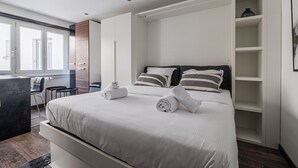 Ein modernes Hotelzimmer mit großem Bett, integriertem Kleiderschrank und einer Sitzecke mit Sessel und Tisch.