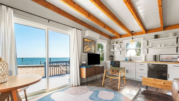 A beachside room with a large sliding glass door to the sea, a kitchen with white cabinets, and a cozy seating area with a brown leather couch. - The Inn Spot (Hampton Bays)
