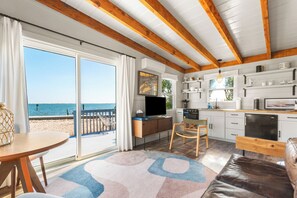 Ein Zimmer am Strand mit einer großen Glasschiebetür zum Meer, einer Küche mit weißen Schränken und einer gemütlichen Sitzecke mit einem braunen Ledersofa.