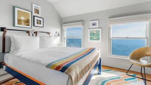 A bedroom with a bed, a chair, and a view of the sea through large windows. - The Inn Spot (Hampton Bays)