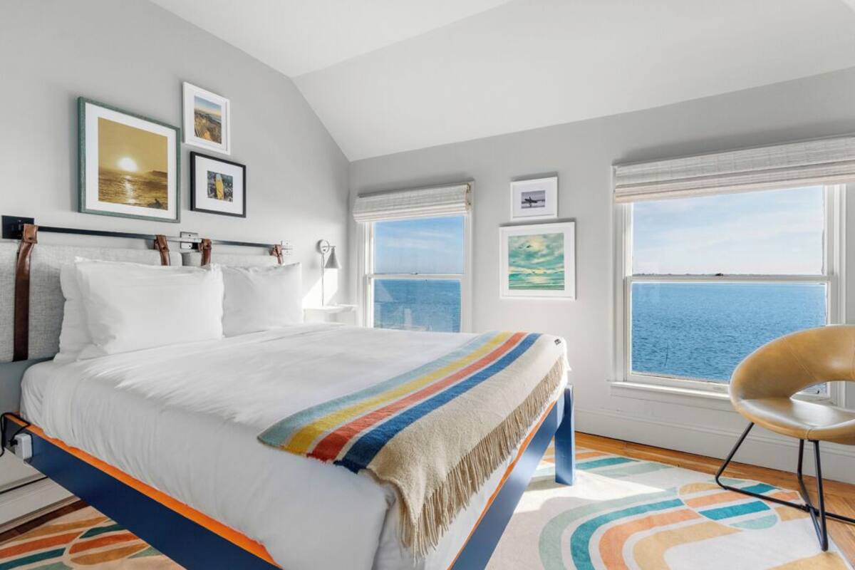 A bedroom with a bed, a chair, and a view of the sea through large windows.