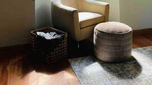 Una habitación en esquina con ventanal saliente, una silla, un pequeño reposapiés y una cesta.