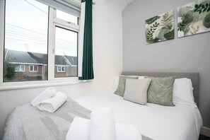 A neatly made bed with pillows and a rolled towel, a window with curtains, and a wall with framed artwork.