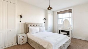 A bedroom with a large bed, bedside tables, a dresser, a vanity with a mirror, and a window with curtains. - Beautiful One-Bedroom Apartment In Notting Hill (London)