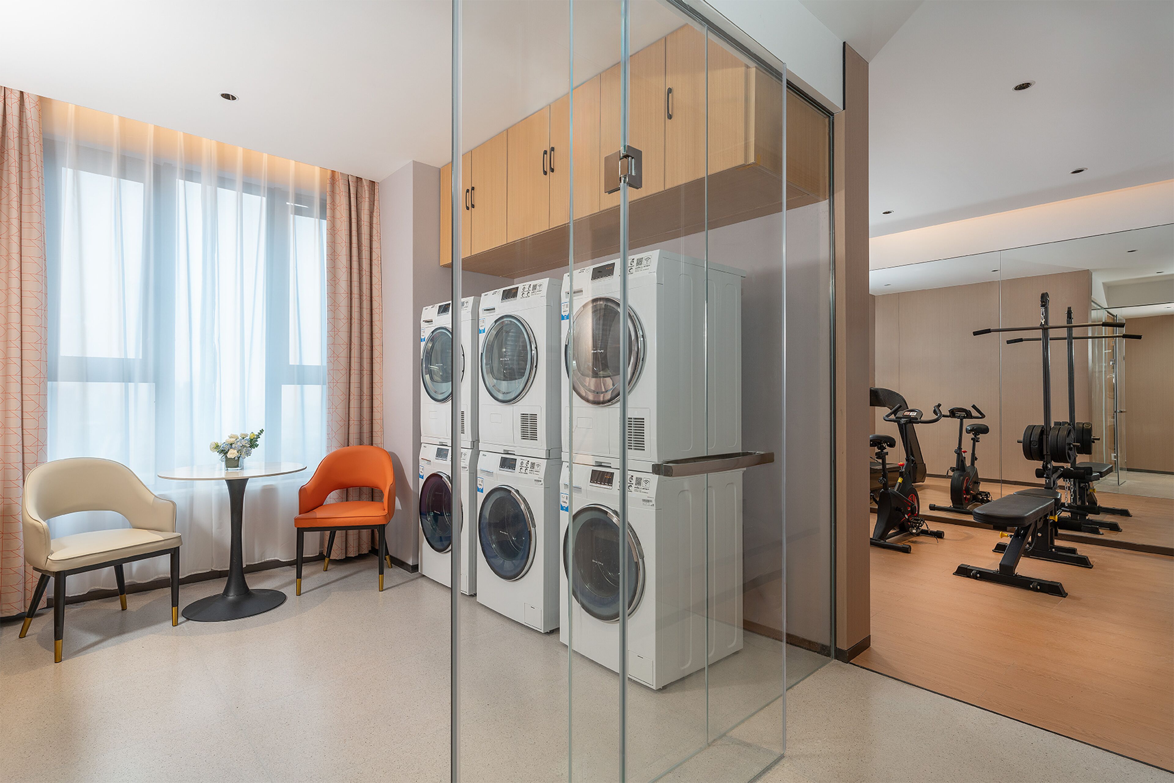 A hotel room with a washing machine, a chair, a table, and a gym area with exercise equipment.