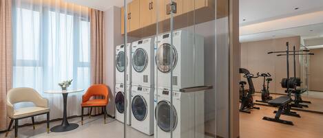 A hotel room with a washing machine, a chair, a table, and a gym area with exercise equipment.