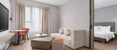 A modern hotel room with a sofa, ottoman, coffee table, and a TV on a desk.