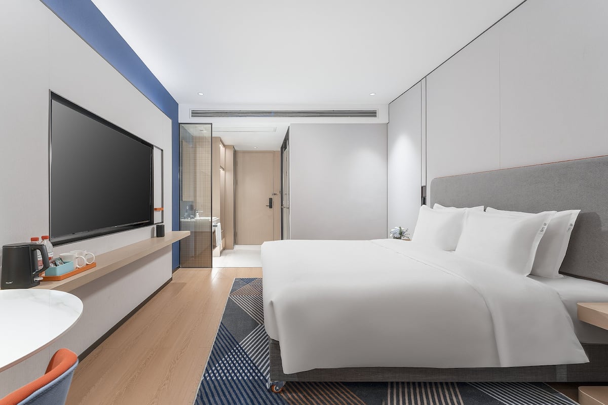 Ein modernes Hotelzimmer mit einem großen Bett, einem Flachbildfernseher, einem Schreibtisch mit Stuhl und einem durch eine Glastür einsehbaren Badezimmer.