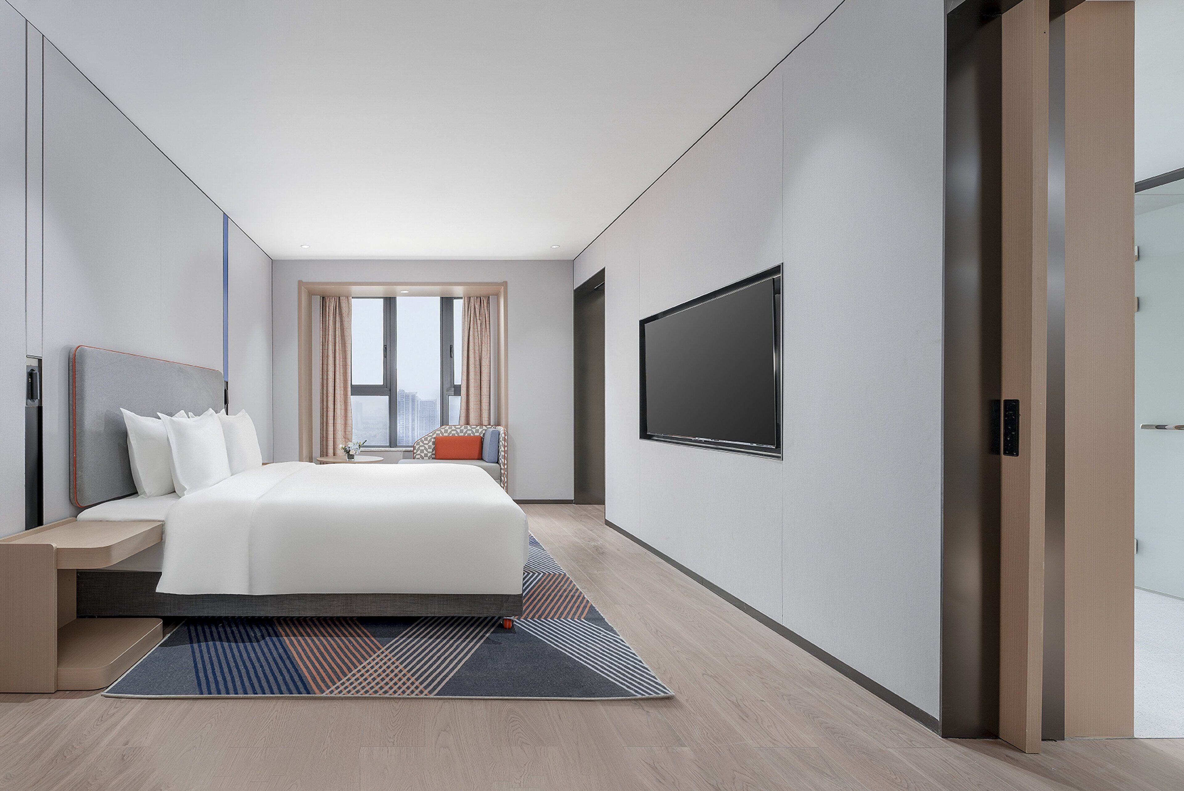 A modern hotel room with a large bed, a flat-screen TV, and a view of the city through the window.