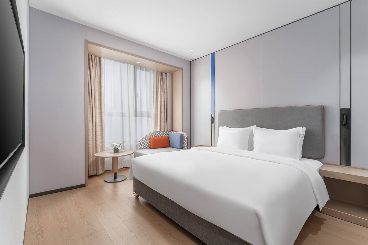 Ein modernes Hotelzimmer mit einem großen Bett, einem kleinen runden Tisch, einem Sessel und einem an der Wand befestigten Fernseher.