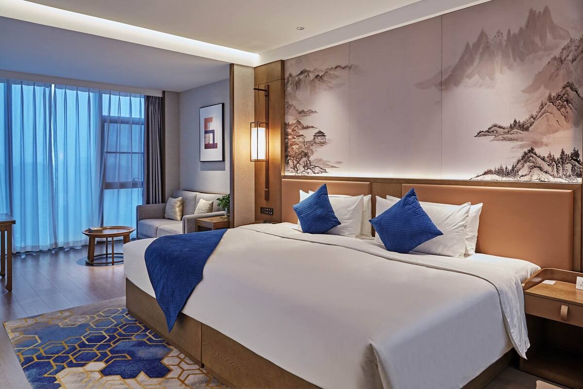 A hotel room with a large bed, a nightstand, a desk, and a chair. There is a window with curtains, a wall-mounted lamp, and a painting of mountains.