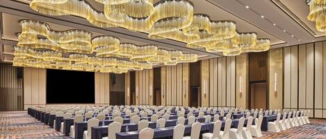 A conference room with rows of tables and chairs, large chandeliers, and a patterned carpet.