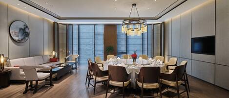 A modern dining area with a round table set for six, surrounded by chairs, in a room with a large window and a television on the wall.