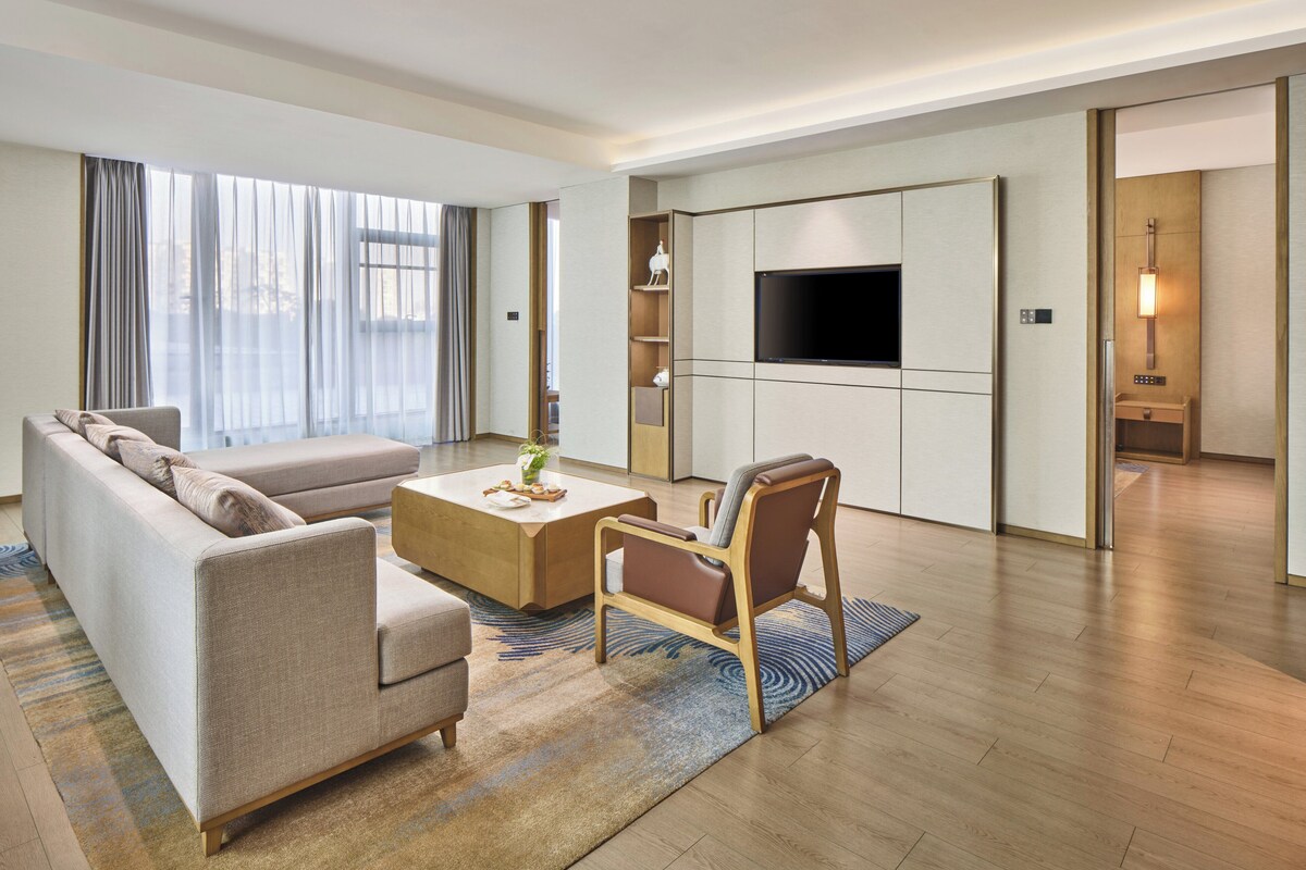 A modern hotel room with a sofa, coffee table, and armchair, a flat-screen TV, and a wooden floor.
