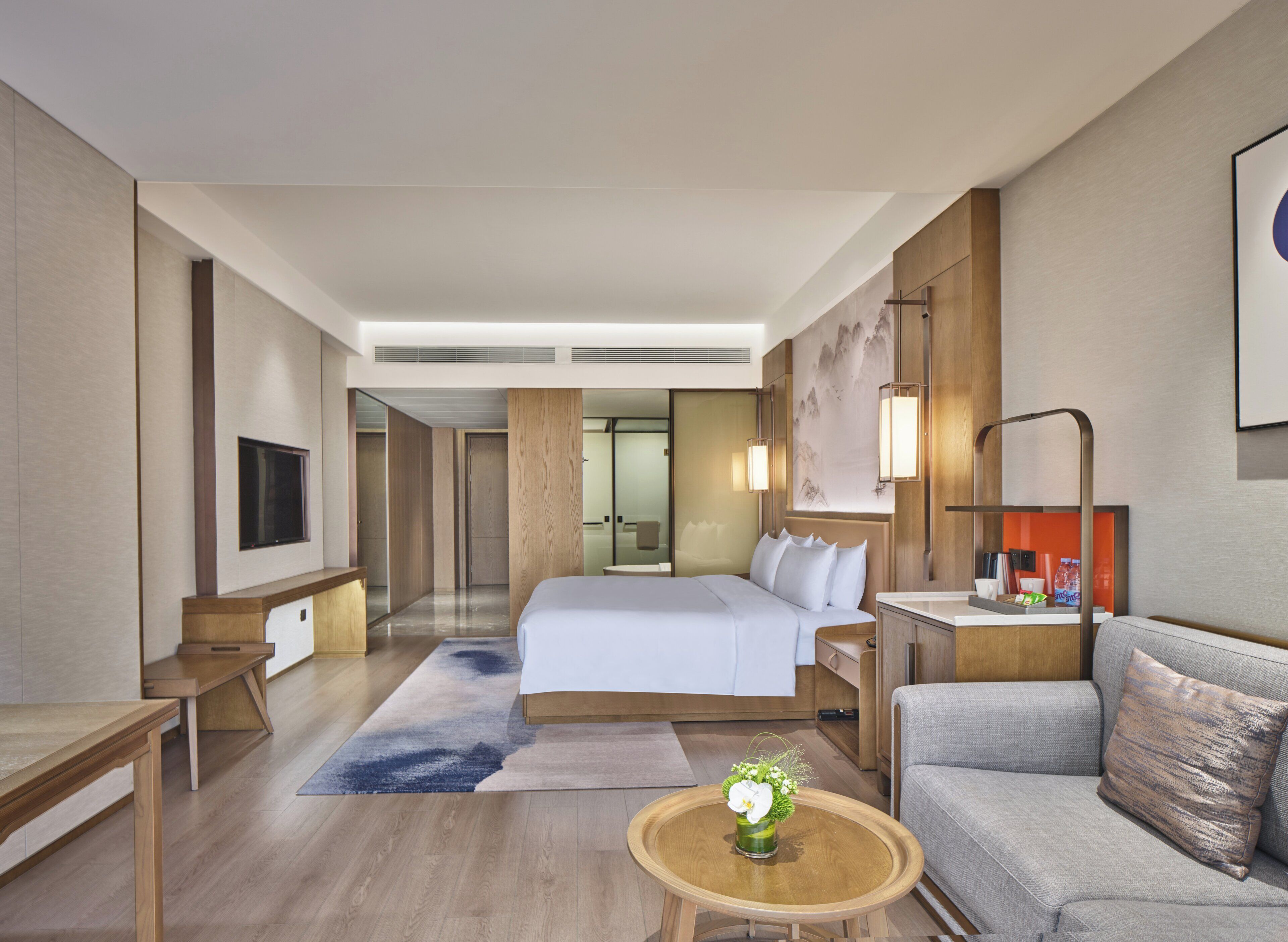 A hotel room with a bed, a sofa, a desk, a TV, and a bathroom visible through a glass door.