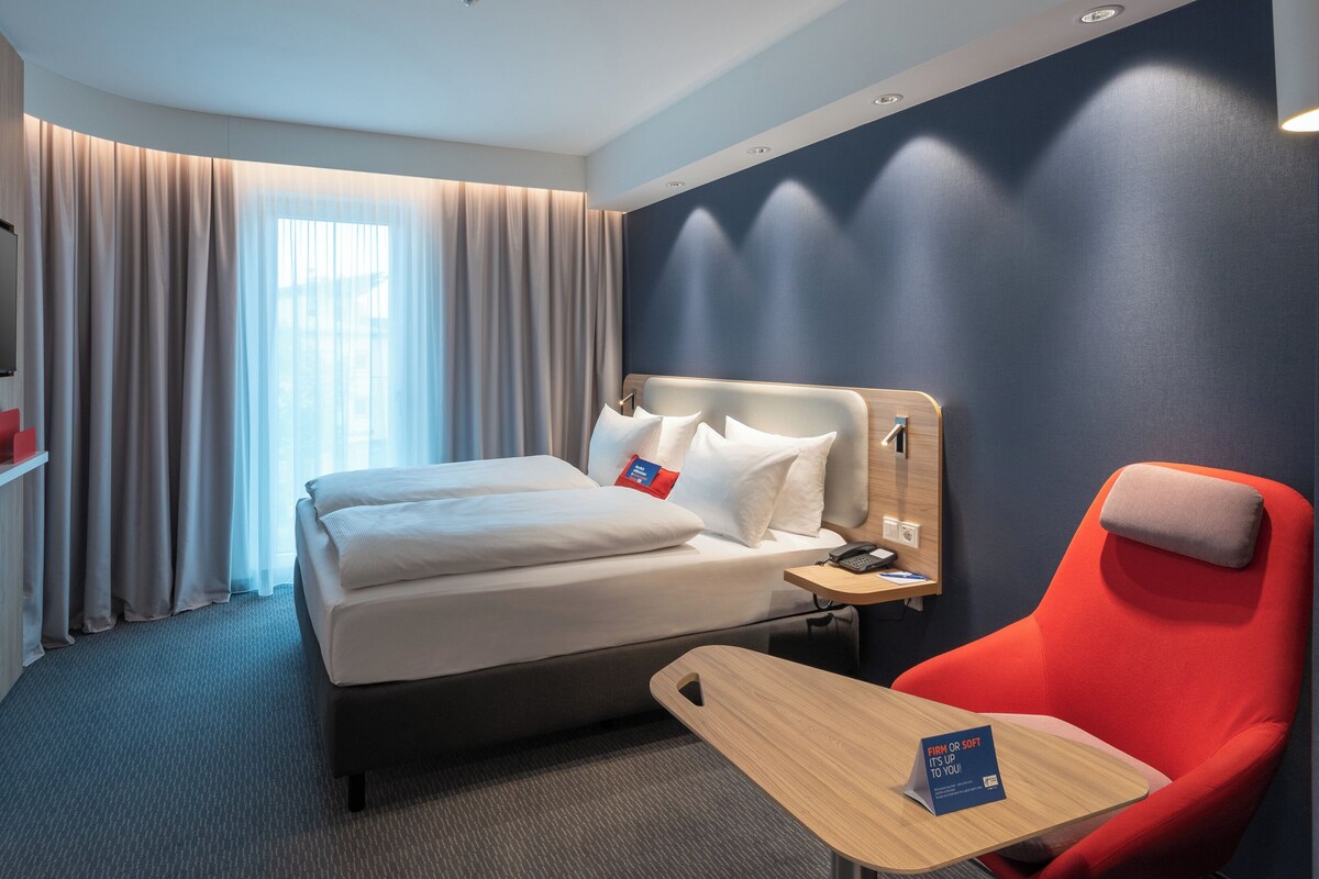 Une chambre d’hôtel avec un lit, un bureau, une chaise, une télévision et des rideaux.