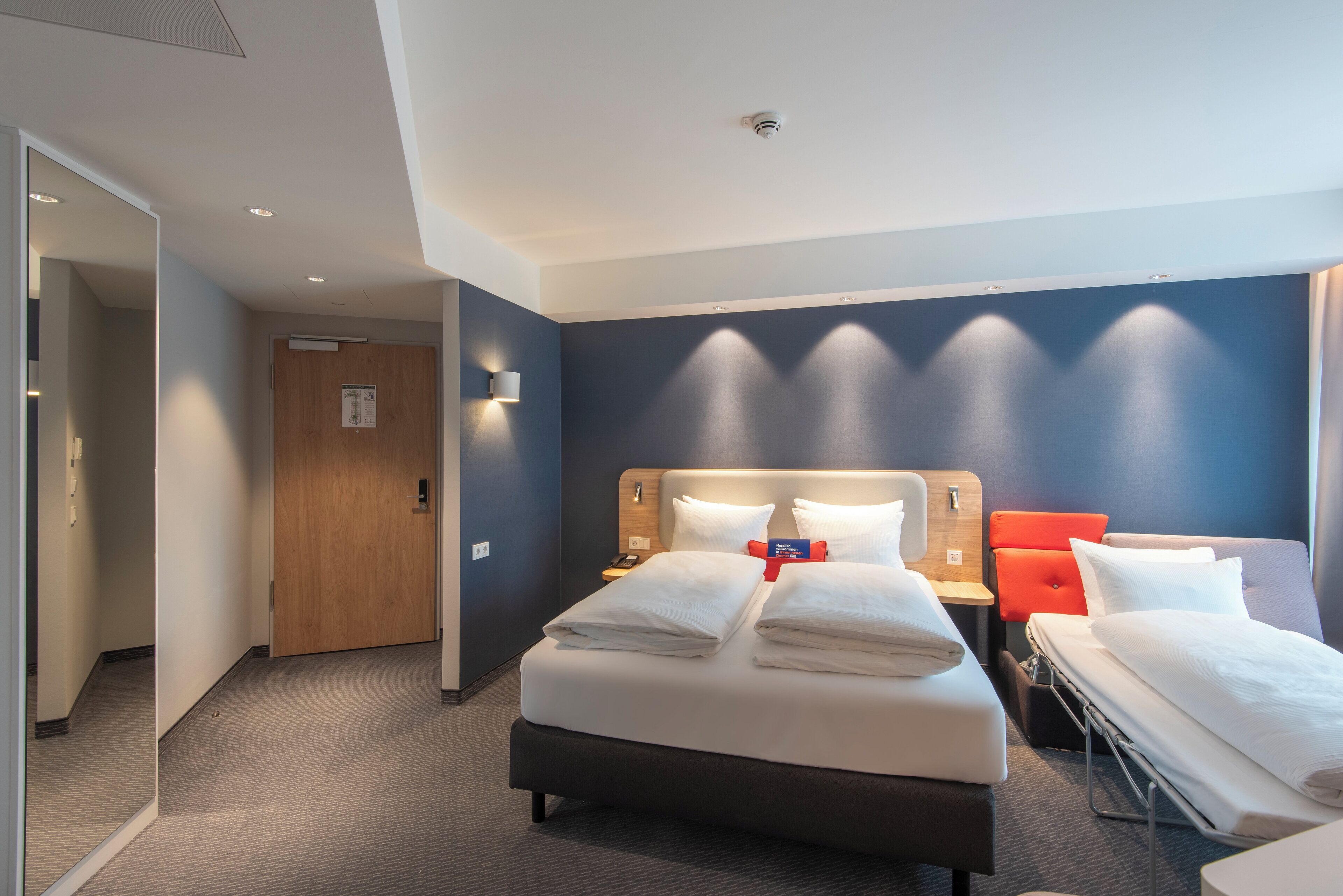 Habitación de hotel con una cama grande, una cama más pequeña, un escritorio y una silla. Hay una puerta de madera y un aplique en la pared.