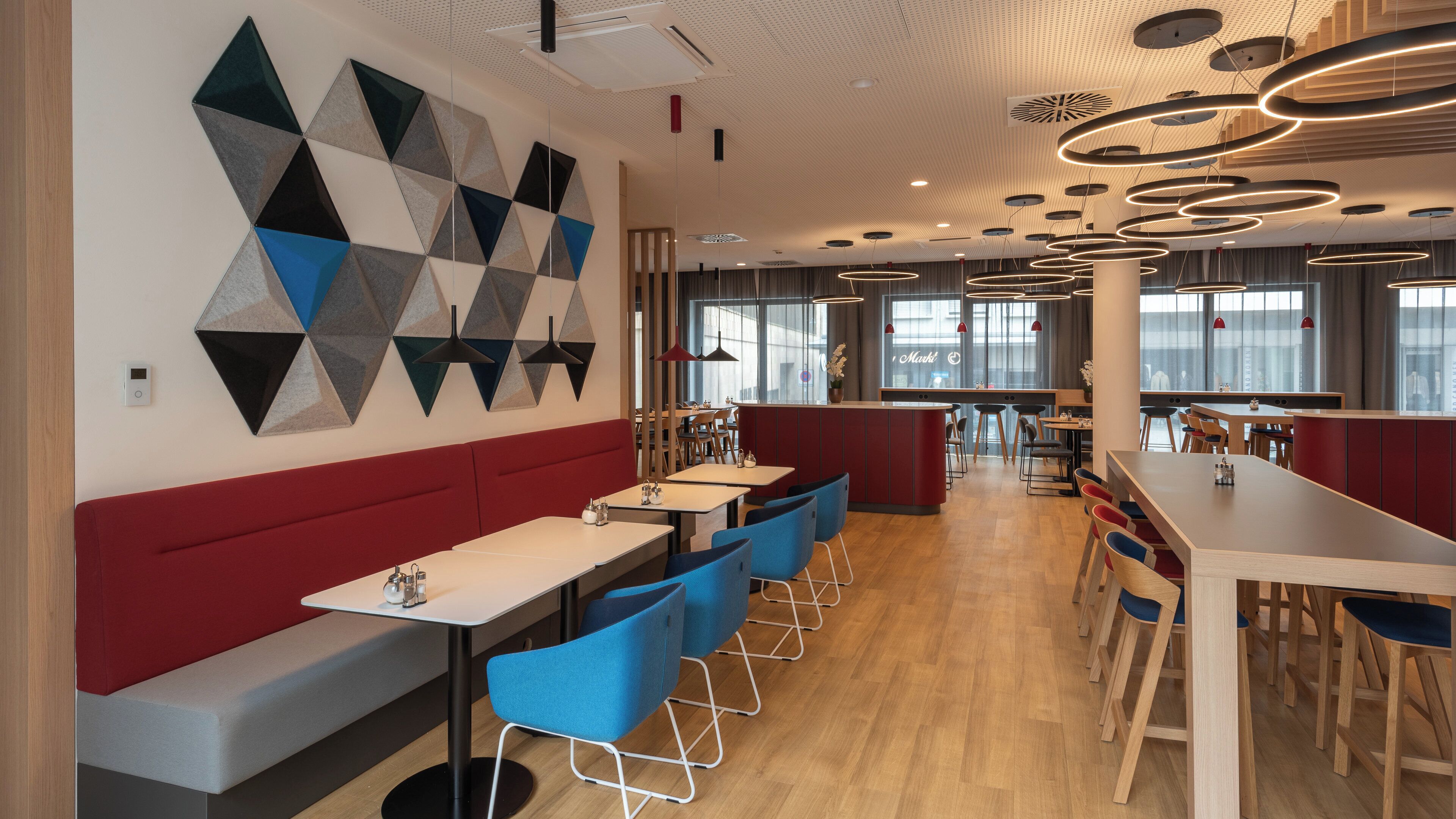 Et moderne restaurantmiljø med geometrisk vægkunst, træborde, blå stole og et rødt siddeområde.