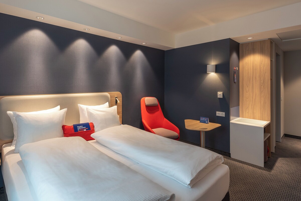 Une chambre d’hôtel avec un grand lit, une chaise rouge, une petite table et une armoire en bois.