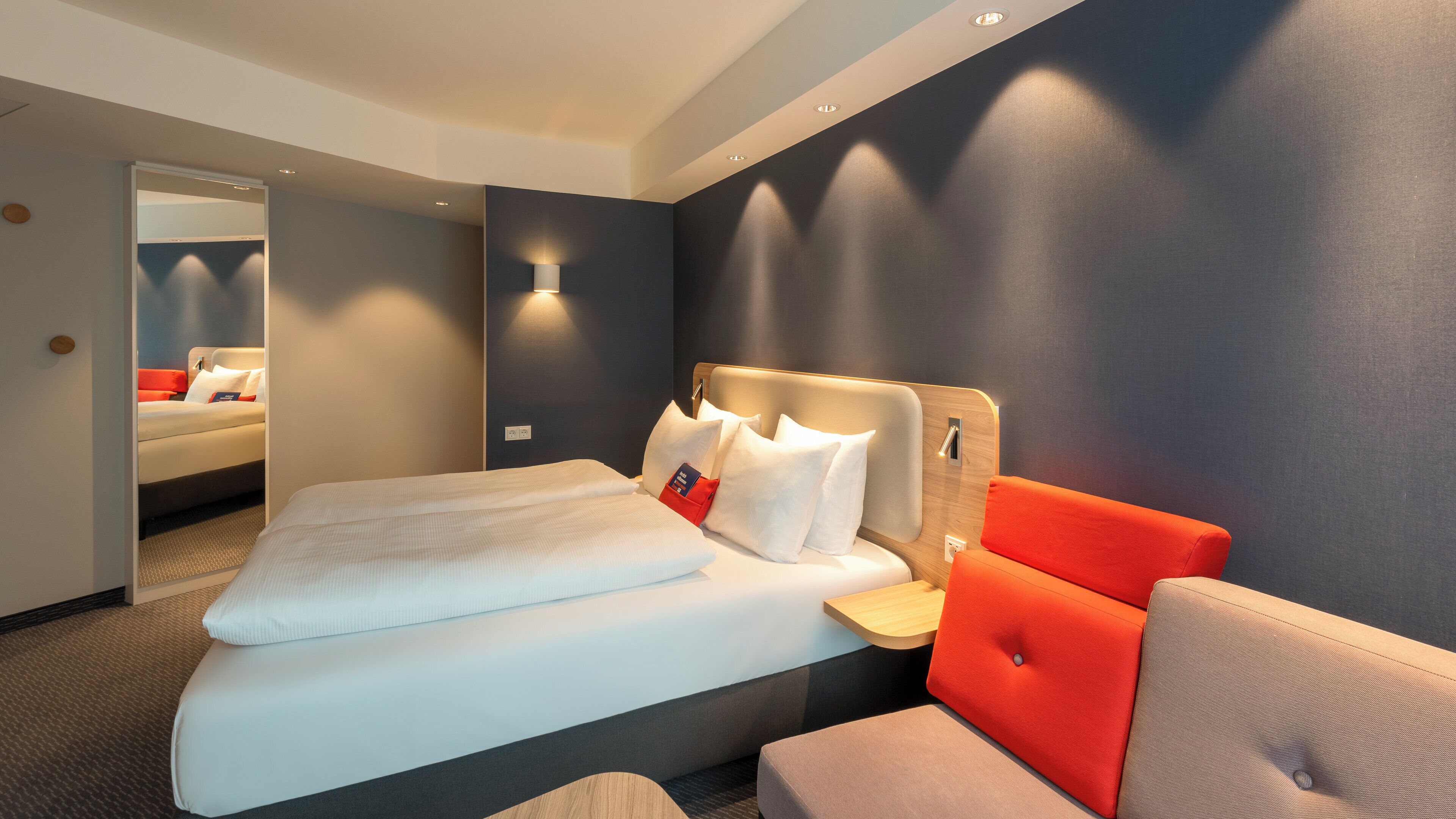 Habitación de hotel con una cama, una mesita de noche, un sillón tapizado rojo y una lámpara de pared.
