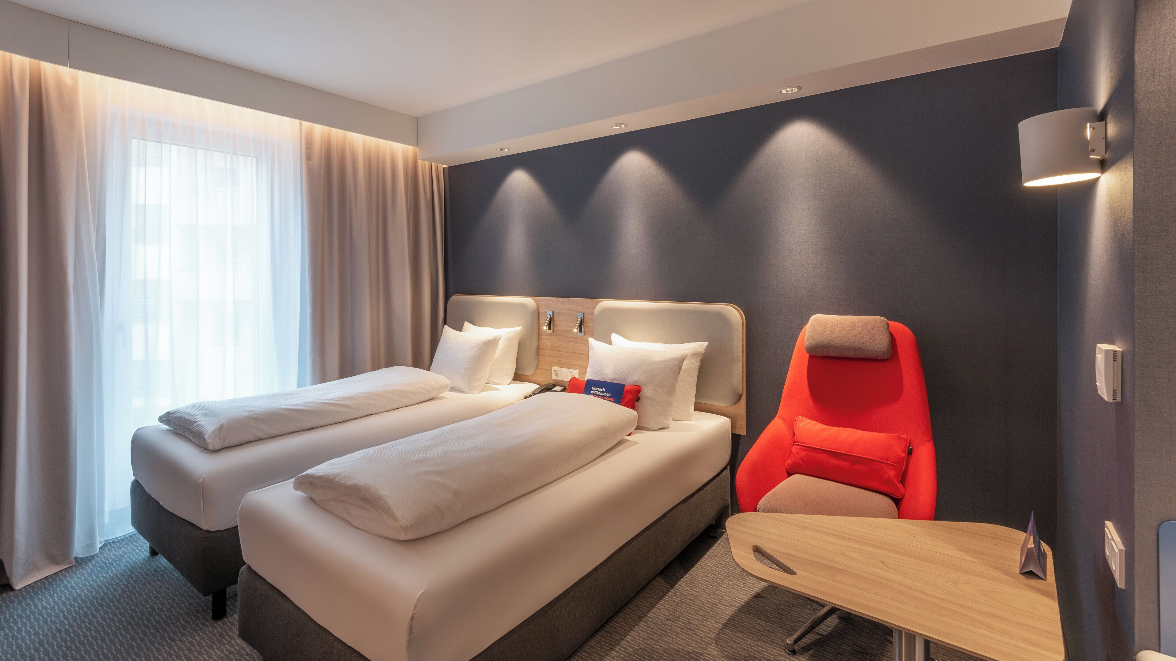 A hotel room with two beds, a red chair, a small table, and a window with curtains.