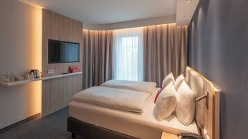 A modern hotel room with a bed, a flat-screen TV mounted on a wooden panel, a bedside table with a kettle and cups, and a window with curtains.