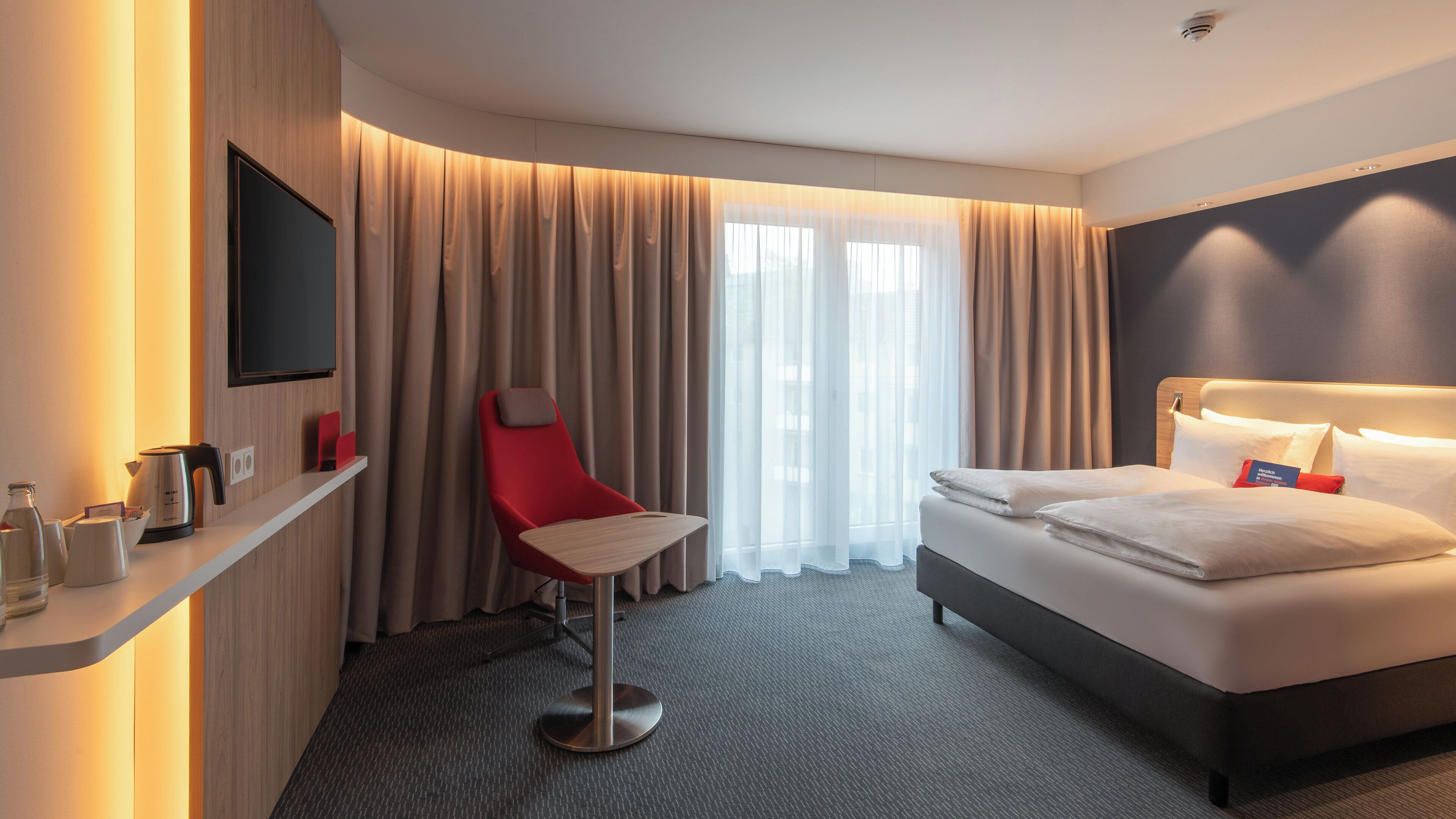 Habitación de hotel con cama, una silla roja, televisión, cafetera y ventana con cortinas.