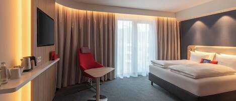 Habitación de hotel con cama, una silla roja, televisión, cafetera y ventana con cortinas.