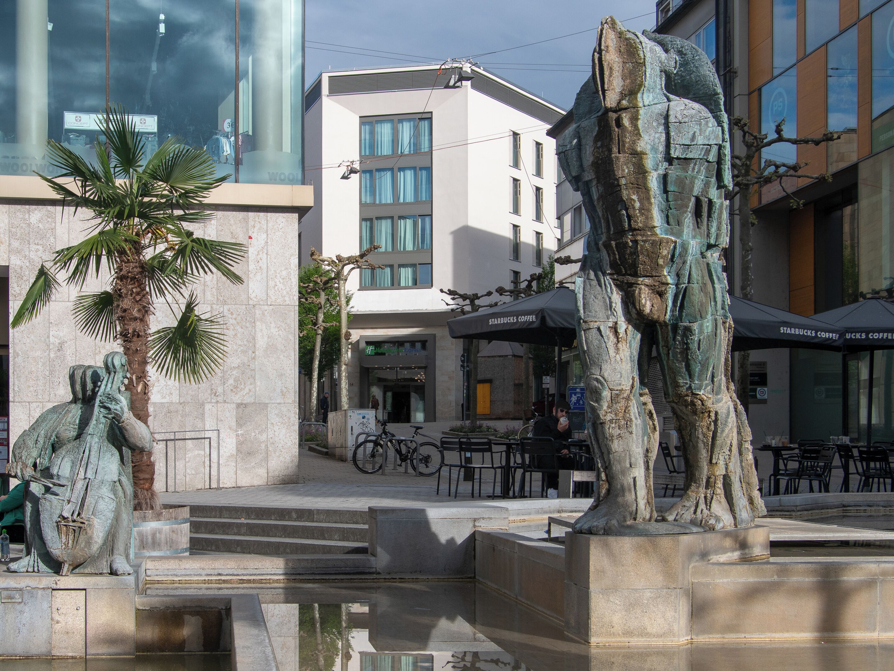 Ein Platz mit einer Bronzestatue, einer Palme und einem Starbucks Café.