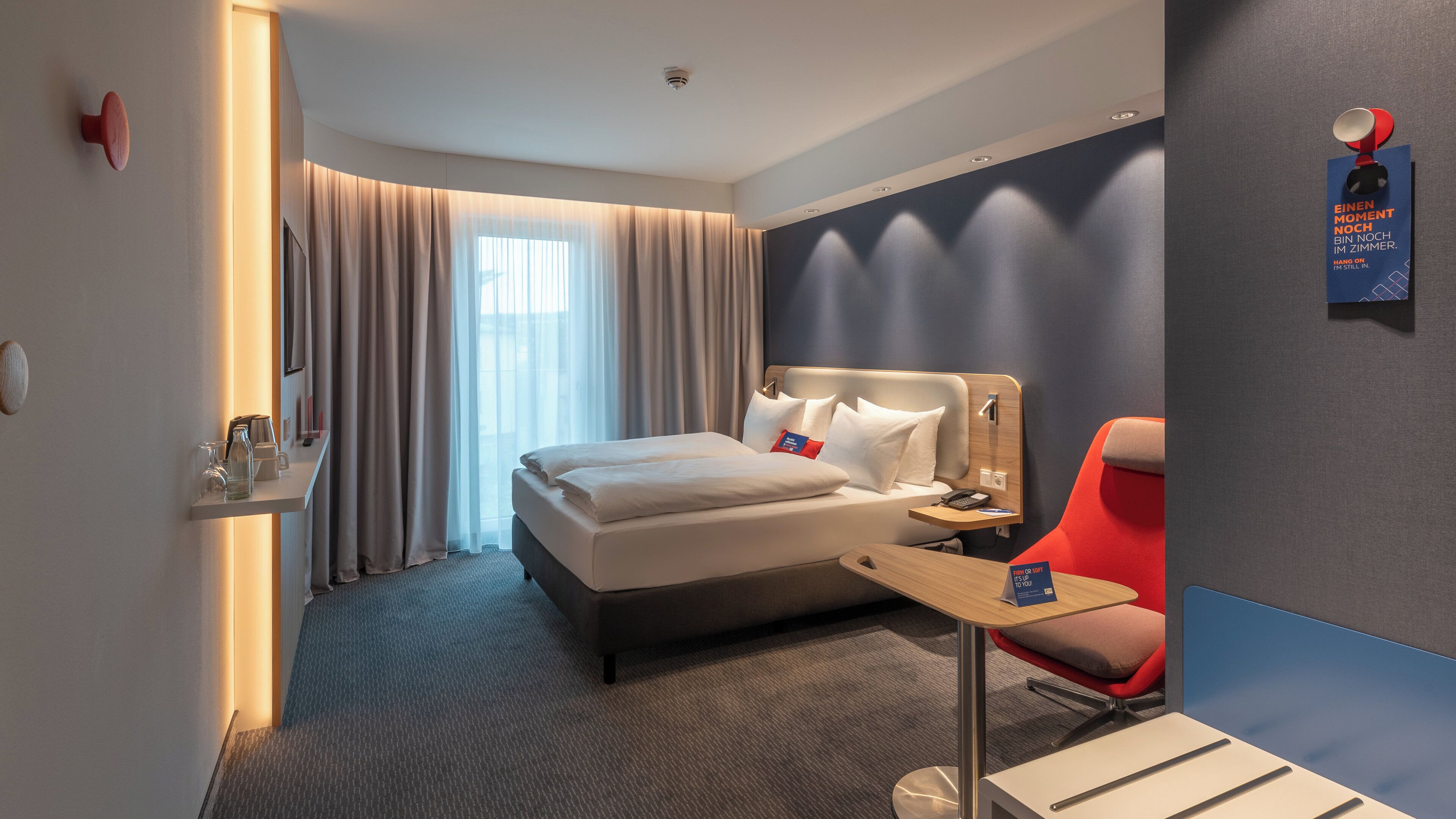 A modern hotel room with a large bed, a bedside table with a lamp, a red chair, and a small table with a sign.