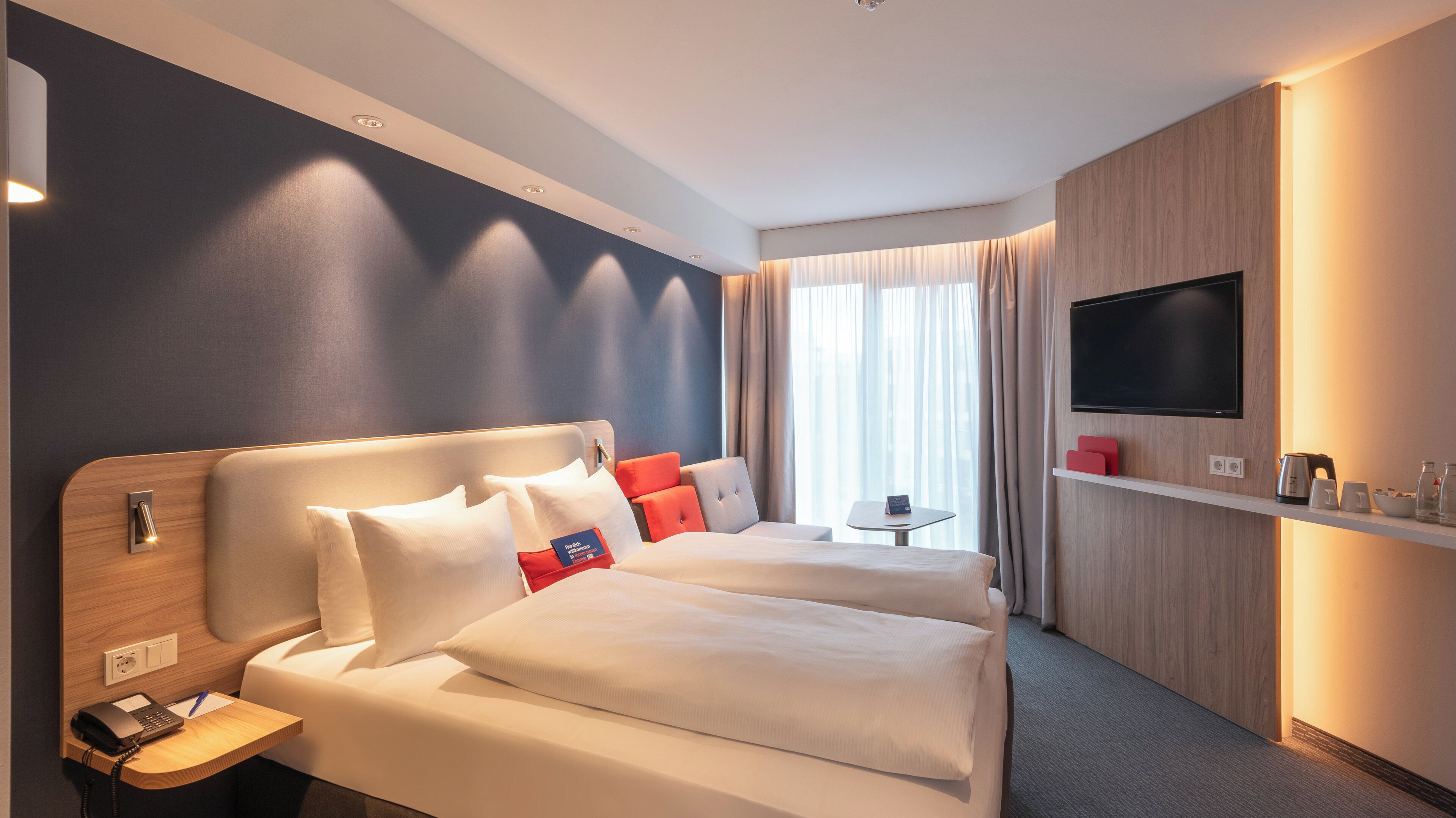 A hotel room with a bed, bedside table, lamp, and a TV on the wall.