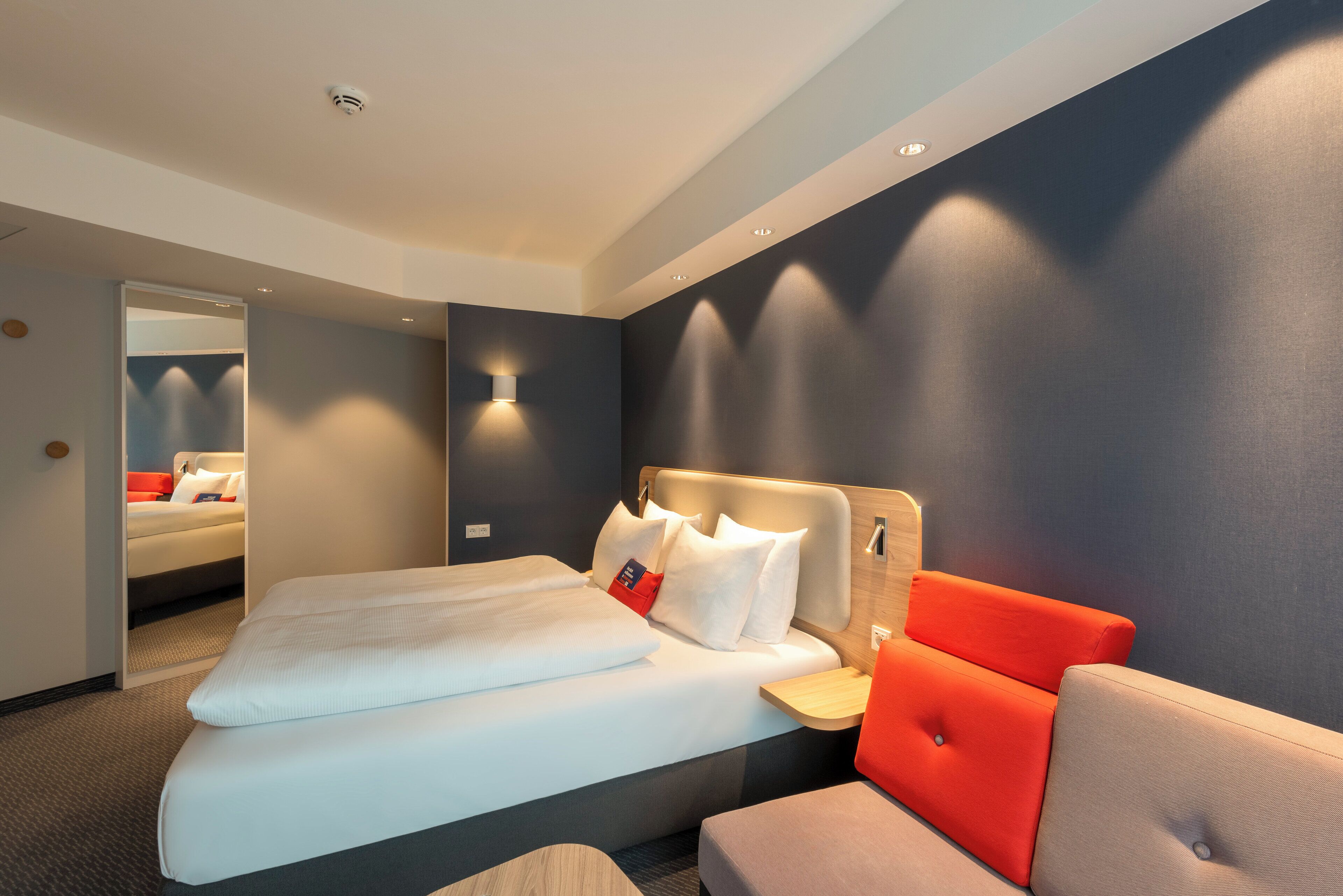 Ein modernes Hotelzimmer mit einem ordentlich bezogenen Bett, einer Sitzecke mit einem rot-grauen Sofa und einer wandmontierten Lampe.