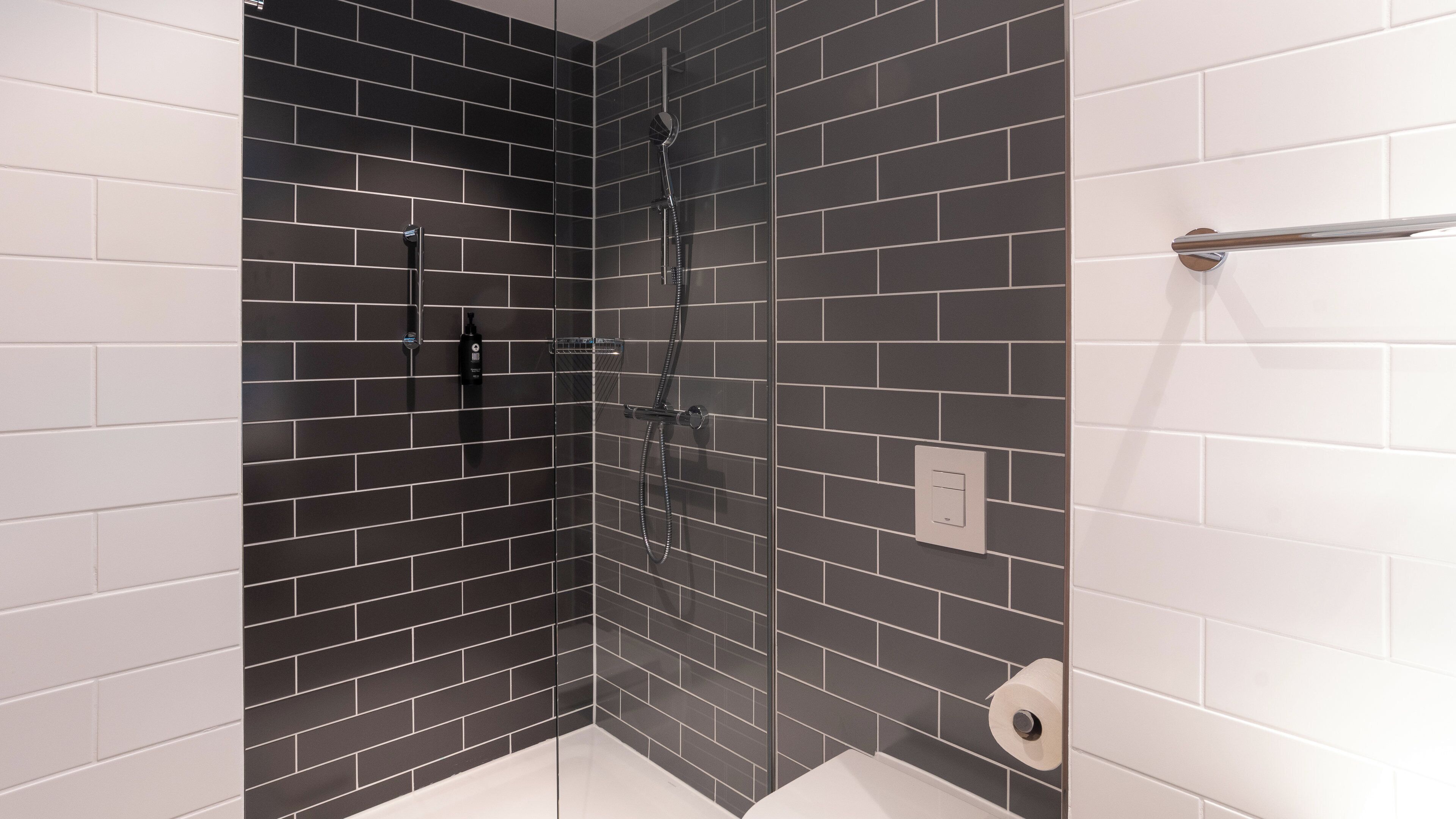 A modern bathroom with dark tiled walls, a glass shower enclosure, and a white tiled floor.