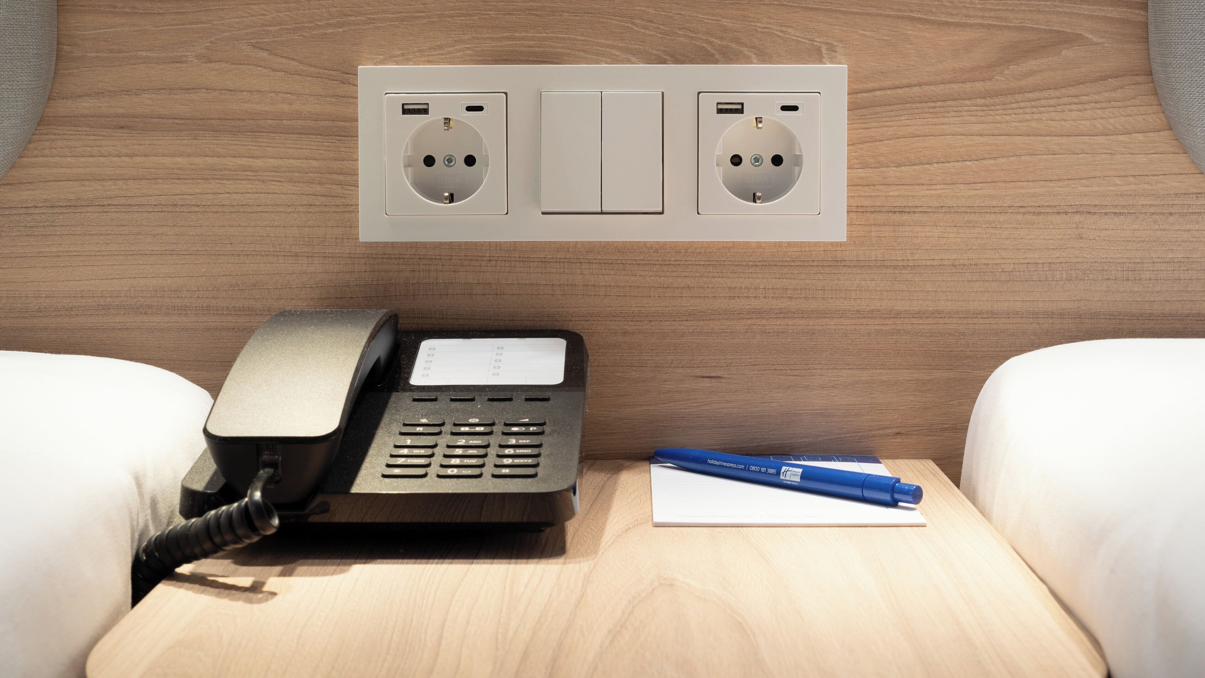 A hotel room with a telephone, a pen, and a notepad on a wooden nightstand.