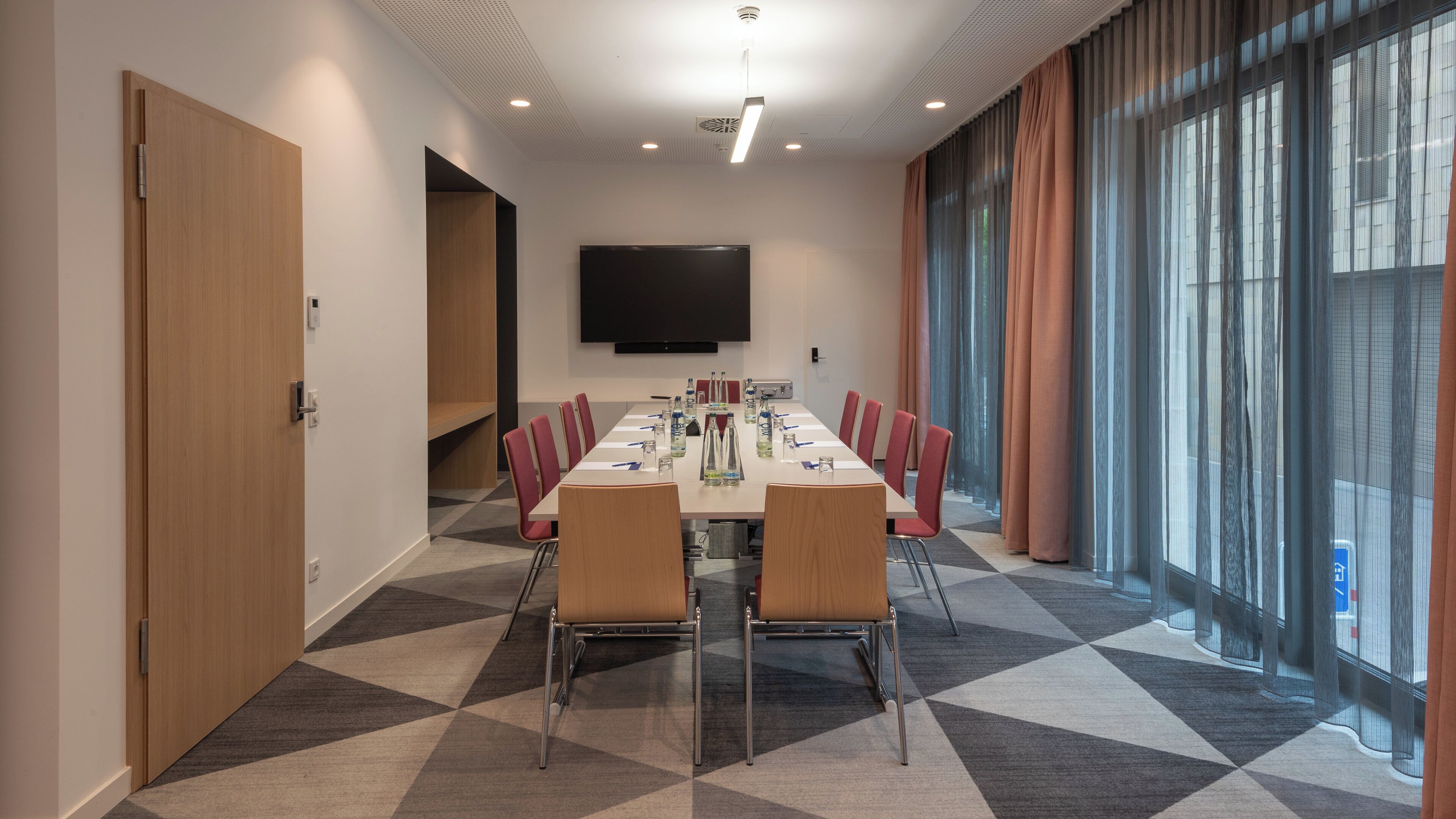 A conference room with a long table, chairs, a flat-screen TV, and large windows with curtains.